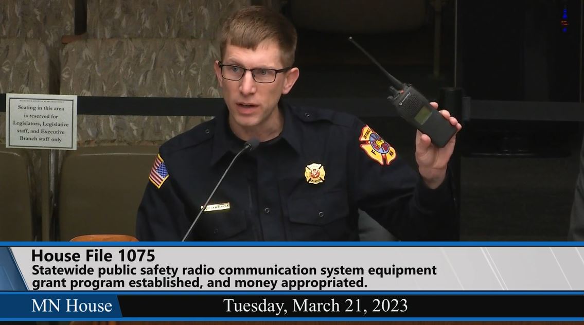 Chuck Henningson, fire chief for the city of Ortonville, holds up a portable radio that became inoperable during a recent emergency call. He testified during Tuesday's House Public Safety Finance and Policy Committee hearing on HF1075. (Screenshot)