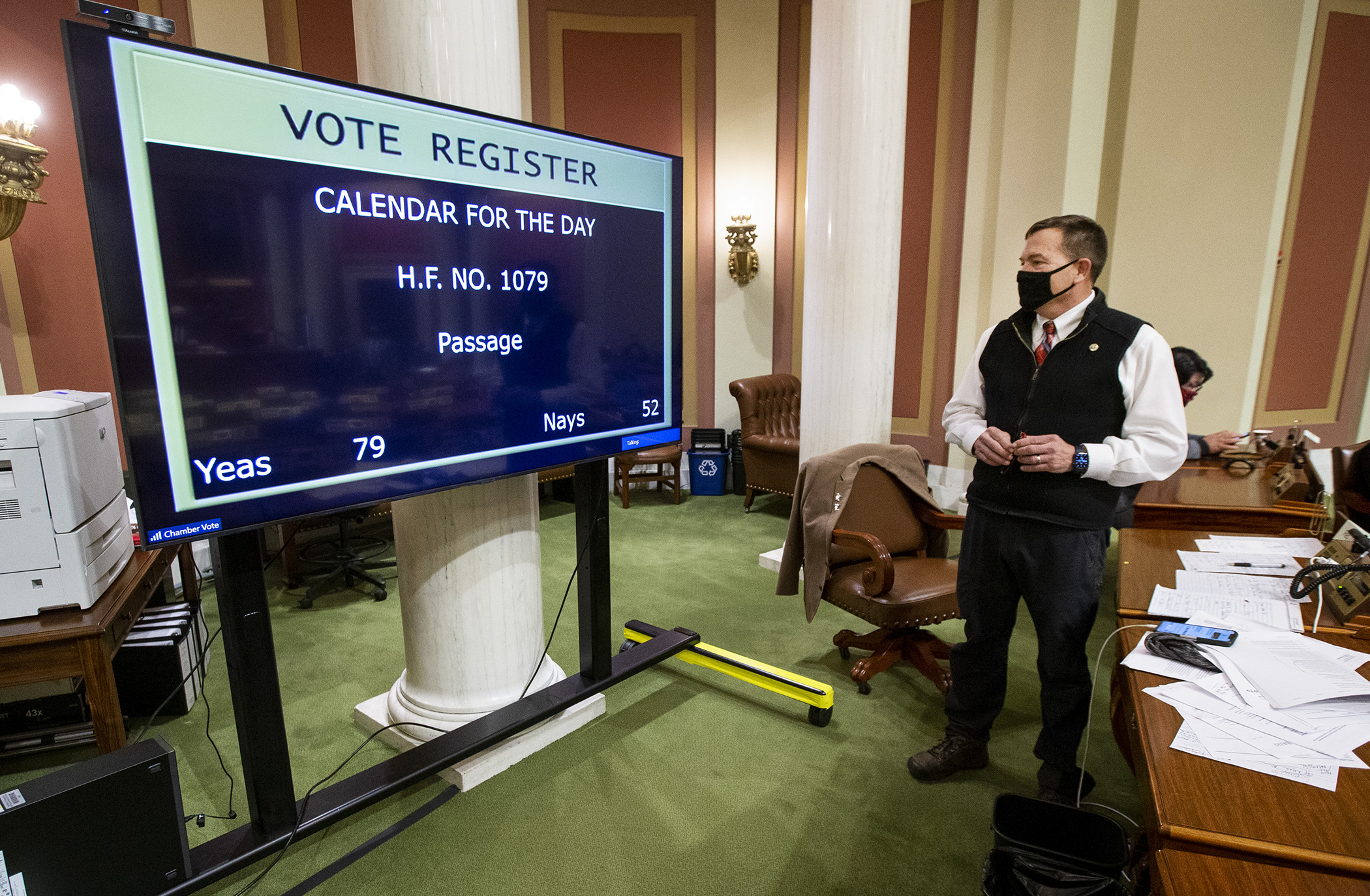 Rep. Leon Lillie, chair of the House Legacy Finance Committee, watches the vote total for the omnibus legacy finance bill on a large TV set up in the back of the House Chamber for visual aid during largely remote floor sessions. Photo by Paul Battaglia