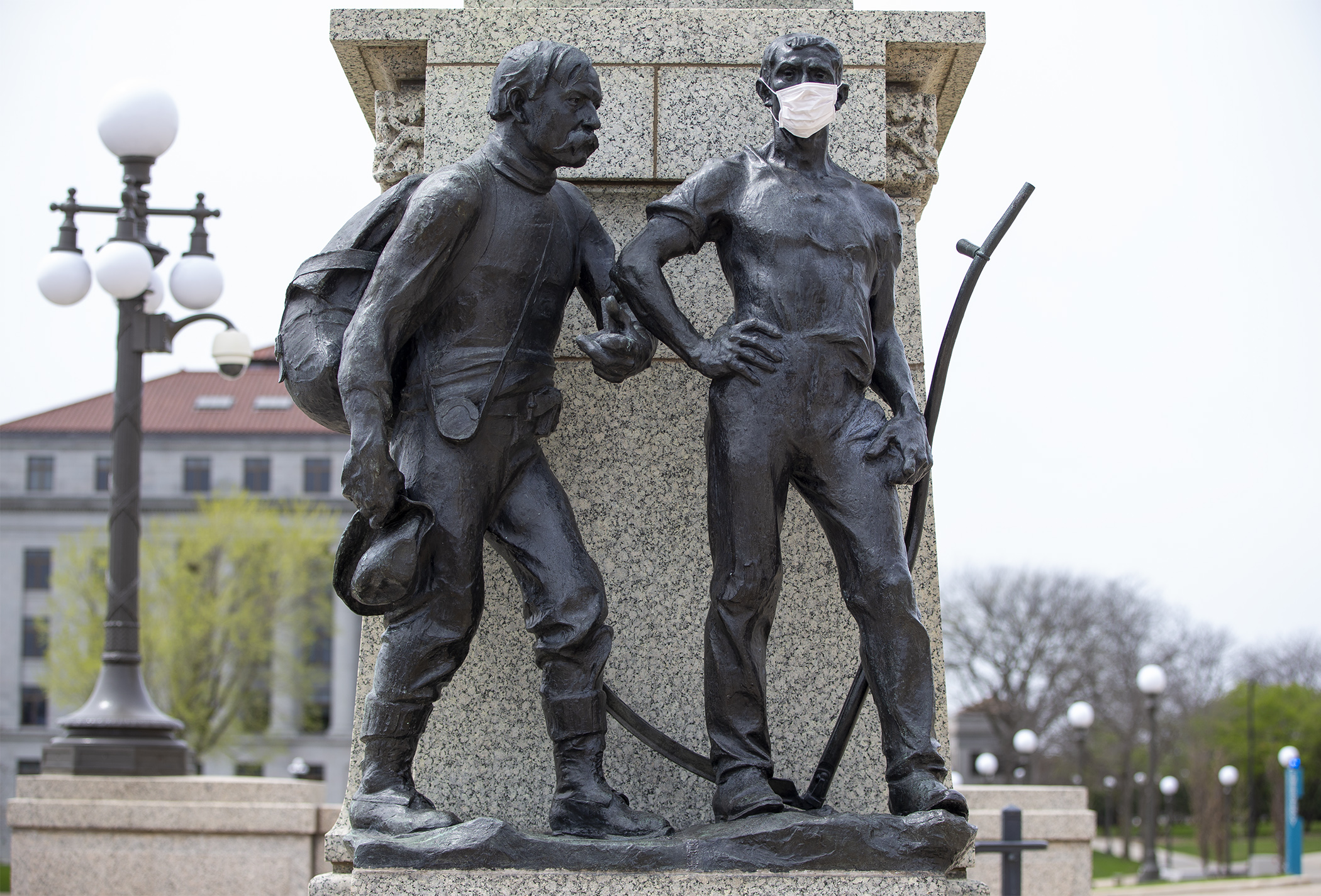 The figure representing agriculture at the base of the Gov. John Johnson statue on the Capitol Mall wears a cloth face covering May 5 as his neighbor, representing manufacturing, disregards social distancing guidelines. Photo by Paul Battaglia